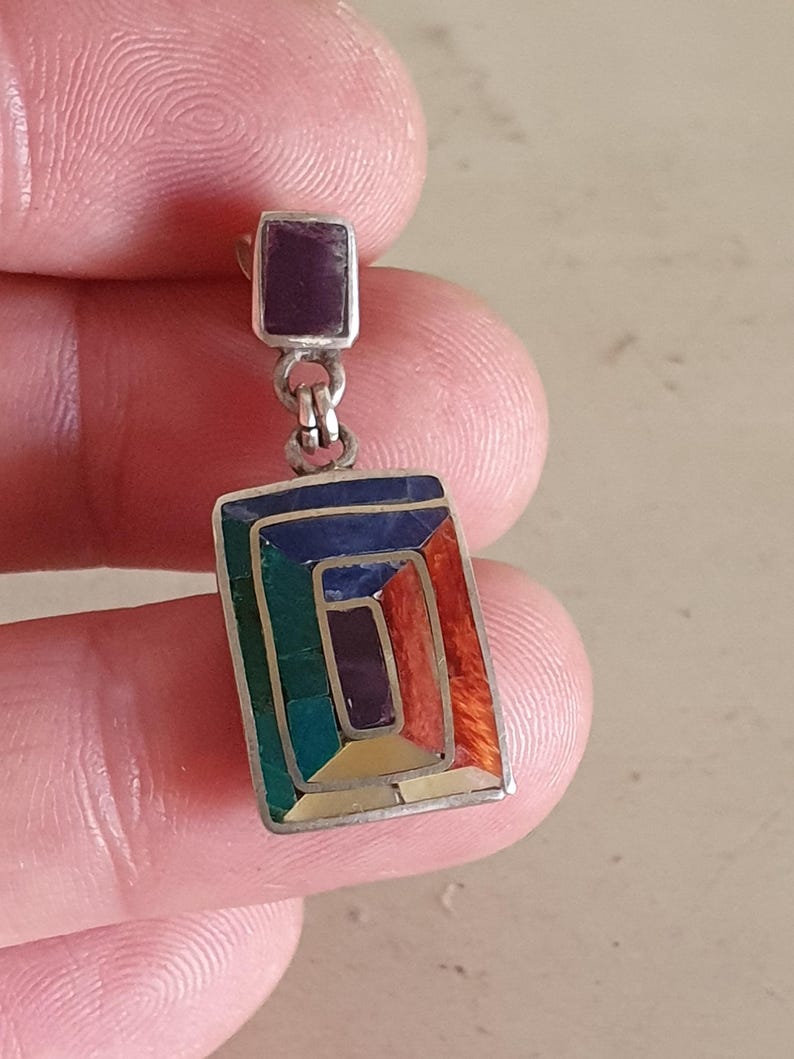 May include: A silver dangle earring with a rectangular shaped stone set in a silver frame. The stone is made of multiple colors of inlaid stones, including green, blue, purple, and orange.