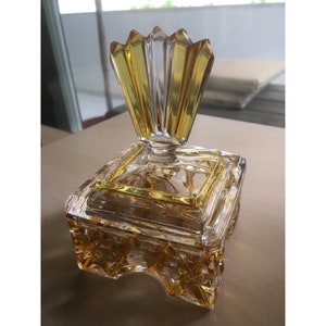Könnte beinhalten: Eine quadratische Glas-Schmuckschatulle mit einem durchsichtigen Deckel und einem gelben, geometrischen Design. Der Deckel hat einen dekorativen, klaren und gelben, fächerförmigen Knauf.