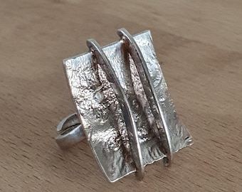 May include: A silver ring with a textured, rectangular face and three parallel, curved bars. The ring has a unique, modern design, suitable for everyday wear or special occasions. The ring is made of a shiny, metallic material.