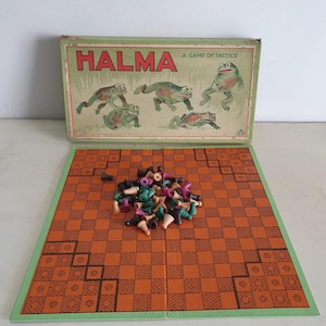 Puede incluir: Juego de mesa vintage Halma con un tablero de ajedrez verde y naranja y una caja con una ilustración verde y marrón de ranas. El juego incluye un conjunto de fichas negras, verdes y moradas.