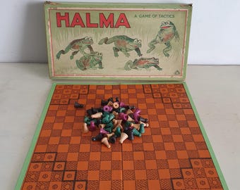 Könnte beinhalten: Vintage Halma Brettspiel mit einem grün-orangen Schachbrett und einer Schachtel mit einer grün-braunen Illustration von Fröschen. Das Spiel enthält einen Satz schwarzer, grüner und violetter Spielsteine.