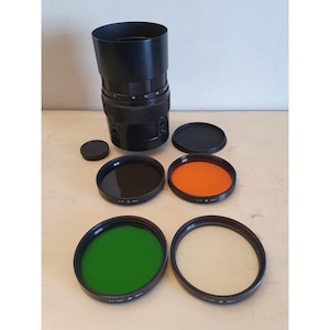 Può includere: Un obiettivo per fotocamera nero con paraluce, tappo dell'obiettivo e quattro filtri colorati: nero, arancione, verde e trasparente. L'obiettivo è cilindrico con indicatori di misurazione. I filtri sono circolari e hanno bordi neri.