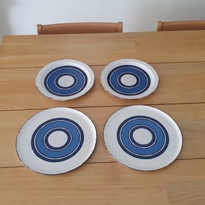 Könnte beinhalten: Vier weiße Keramikplatten mit einem blauen konzentrischen Kreismuster. Die Teller sind auf einem hellen Holztisch angeordnet. Die Teller haben einen Durchmesser von etwa 23 cm.