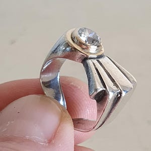 May include: A silver ring with a gold band holding a clear, round gemstone. The ring has a unique geometric design with a fan-like shape on the side. The ring is held by a person's fingers.