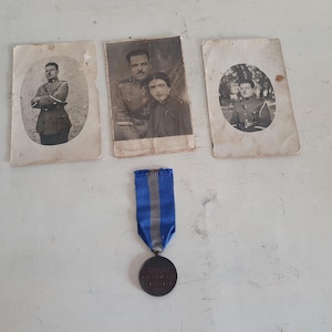 Peut inclure: Trois photographies anciennes en noir et blanc de personnes en uniforme militaire. Les photos sont dans des cadres rectangulaires et sont montées sur du carton blanc. Un ruban bleu et blanc avec une médaille de bronze est au centre de l'image.