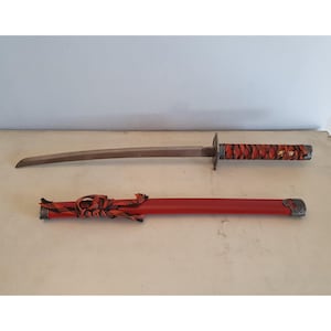 Könnte beinhalten: Ein japanisch anmutendes Schwert mit gebogener Klinge und einem rot-schwarz umwickelten Griff. Das Schwert wird mit seiner roten Scheide präsentiert, die silberfarbene Akzente und einen dekorativen Knoten aufweist.