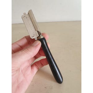 May include: A vintage metal and wood vegetable peeler with a black wooden handle. The peeler has a silver blade and a small metal piece with text. The handle is smooth and rounded.