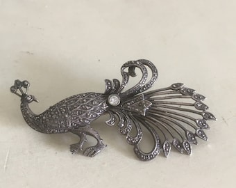 May include: A vintage silver-toned peacock brooch. The brooch features intricate detailing, including a central clear gemstone and a fanned tail. The peacock is depicted in a side profile, with its head and body detailed with small, textured accents.