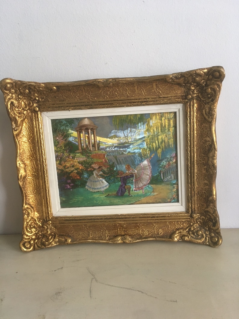 Op de afbeelding: Een ingelijst schilderij dat een sc&egrave;ne met drie figuren in een tuinsetting toont. De figuren zijn gekleed in 18e-eeuwse kleding en zijn bezig met een dans of een uitvoering. De achtergrond toont een waterval, een prieel en weelderig groen.