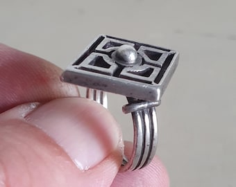 May include: A silver ring with a square-shaped top featuring a geometric design. The ring has a twisted band.