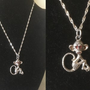 May include: A silver chain necklace with a silver monkey pendant. The monkey has red eyes and is sitting with its tail curled around its legs.