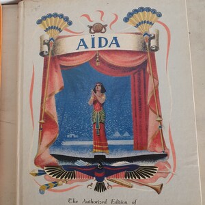 May include: A vintage book cover featuring a colorful illustration of a woman in a red dress standing on a stage with the title "AIDA" in a banner above her. The book is titled "The Authorized Edition of THE METROPOLITAN OPERA GUILD, INC."