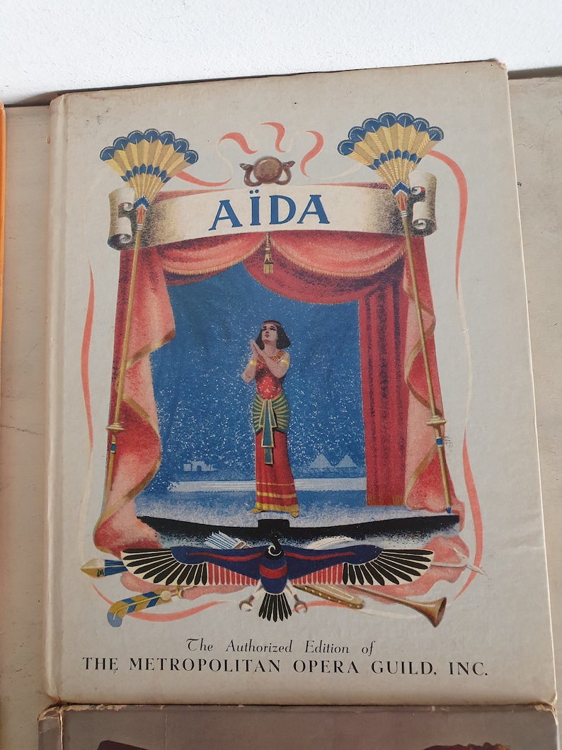 May include: A vintage book cover featuring a colorful illustration of a woman in a red dress standing on a stage with the title "AIDA" in a banner above her. The book is titled "The Authorized Edition of THE METROPOLITAN OPERA GUILD, INC."