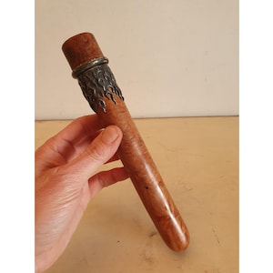 Könnte beinhalten: Ein Zigarrenhalter aus Holz mit einem dekorativen Metallband. Der Halter hat eine hellbraune Farbe mit einer dunkleren braunen Kappe. Das Metallband hat ein flammenartiges Design. Der Zigarrenhalter wird in der Hand einer Person gehalten.