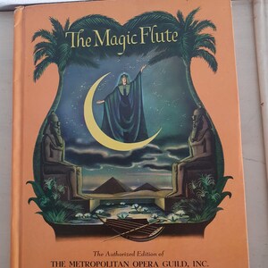 May include: The Magic Flute book cover with an illustration of a woman in a green robe standing on a crescent moon with pyramids in the background. The text "The Authorized Edition of THE METROPOLITAN OPERA GUILD, INC." is at the bottom of the cover.
