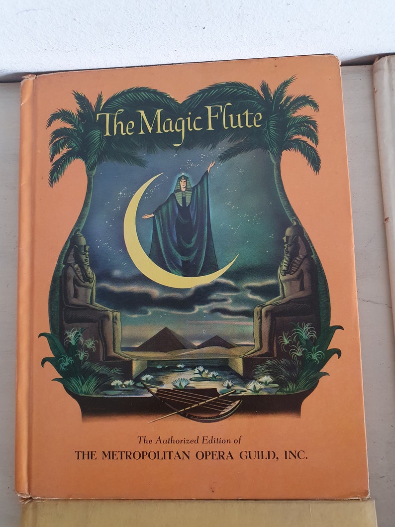 May include: The Magic Flute book cover with an illustration of a woman in a green robe standing on a crescent moon with pyramids in the background. The text "The Authorized Edition of THE METROPOLITAN OPERA GUILD, INC." is at the bottom of the cover.
