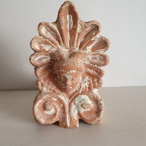Peut inclure: Une sculpture antique en terre cuite de couleur terracotta représentant un visage stylisé avec une coiffe. La sculpture présente un design détaillé avec un aspect vieilli, montrant des traces de peinture blanche et bleue. Il s'agit probablement d'un élément décoratif.