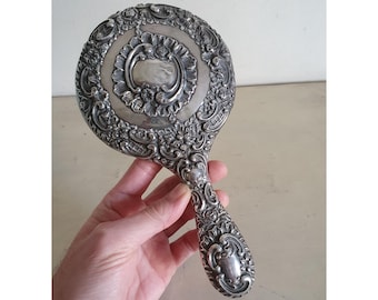 May include: A silver hand mirror with an ornate, floral design. The mirror has a round face and a long, detailed handle.