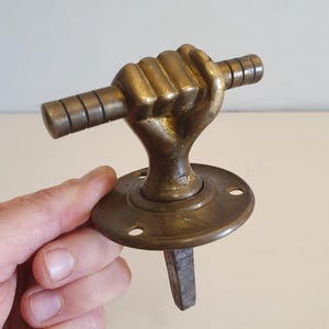 Peut inclure: Poignée de porte ancienne en laiton représentant une main serrée tenant une barre horizontale. La poignée a une base circulaire avec des trous de vis et une tige carrée. Le laiton présente une patine chaude et vieillie.