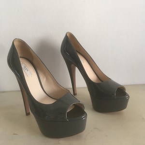 Pode incluir: Um par de sapatos de plataforma de couro patenteado preto com bico aberto. Os saltos medem aproximadamente 13 cm de altura e a plataforma cerca de 2,5 cm. Os sapatos são da Prada.