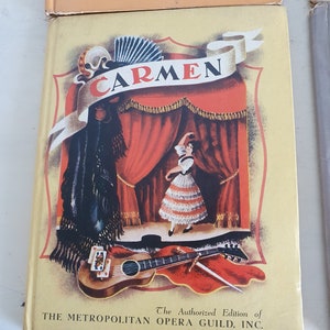 May include: A vintage book cover featuring a colorful illustration of a woman in a red and white dress performing on stage. The title "CARMEN" is displayed in a banner above the stage. The book is an authorized edition of The Metropolitan Opera Guild, Inc.