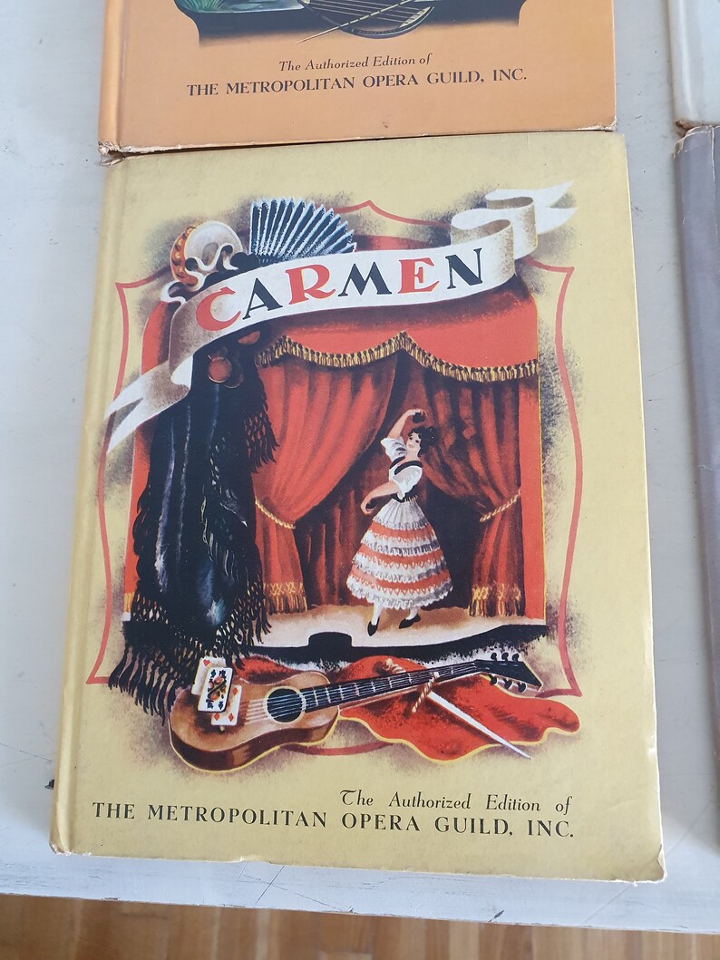 May include: A vintage book cover featuring a colorful illustration of a woman in a red and white dress performing on stage. The title "CARMEN" is displayed in a banner above the stage. The book is an authorized edition of The Metropolitan Opera Guild, Inc.