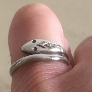 Peut inclure: Une bague argentée en forme de serpent enroulé autour d'un doigt. La tête du serpent présente deux petits détails sombres pour les yeux et un motif texturé. La bague est un bijou.