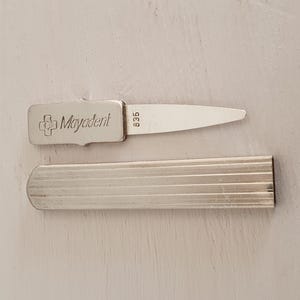 May include: A silver dental instrument with the text "Mayadent" and a cross symbol. The instrument has a blade and a handle, along with a separate silver case. The number "835" is also visible on the instrument.