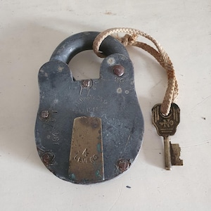 Puede incluir: Un candado vintage de color gris oscuro con una placa de latón que dice "4 LEVERS" y "1847" en la parte inferior. El candado tiene una cerradura y una llave atada a una cuerda marrón.