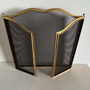 May include: A three-panel fireplace screen featuring a gold-toned metal frame with a curved top edge and a fine black mesh screen. The two side panels are hinged to the central panel, creating a foldable structure with a metallic finish.
