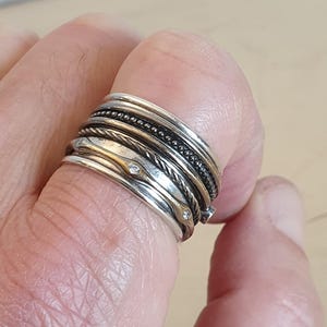 May include: A multi-layered silver ring with various textures and designs. The ring features several bands, including twisted, beaded, and smooth surfaces. Small, clear gemstones are set into some of the bands. The ring is worn on a finger.