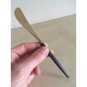 May include: A gold butter knife with a black handle. The knife has the word "Caywood" engraved on the blade.