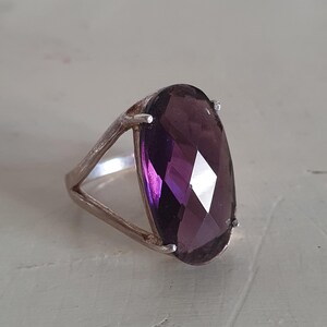 Könnte beinhalten: Ein Silberring mit einem großen, facettierten, oval geschliffenen Amethyst-Edelstein. Der tiefviolette Stein ist in einem schlichten Silberband mit geteiltem Schaft gefasst. Der Edelstein reflektiert das Licht.