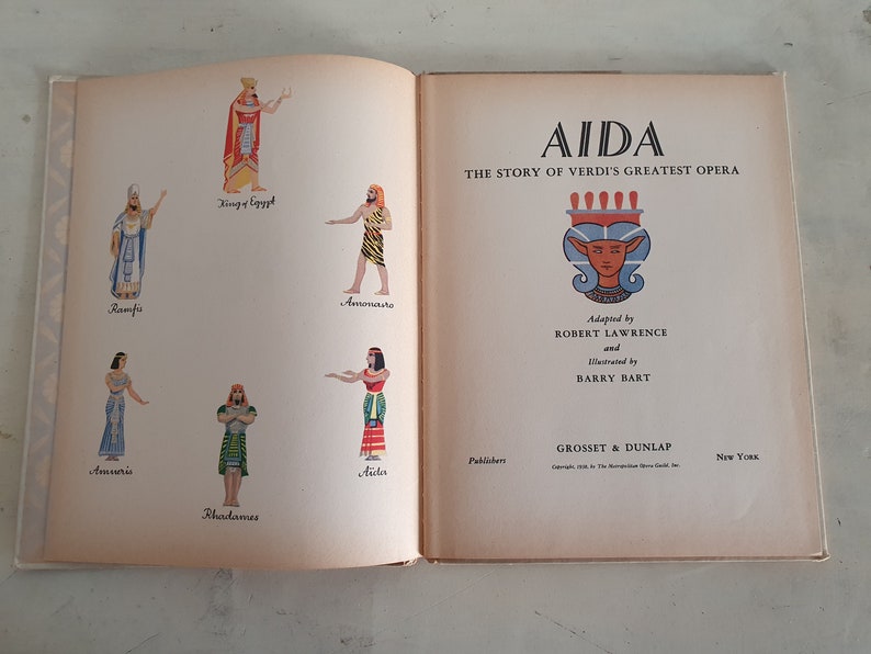 May include: A book cover with the title "AIDA THE STORY OF VERDI'S GREATEST OPERA" and illustrations of characters from the opera. The cover is beige with a red and gold design.