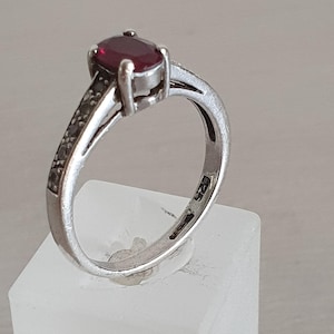 May include: A silver ring featuring a central oval-cut deep red gemstone held in a four-prong setting. The band is crafted from polished silver and includes the hallmark 925 stamped on the inner surface, indicating sterling silver composition.