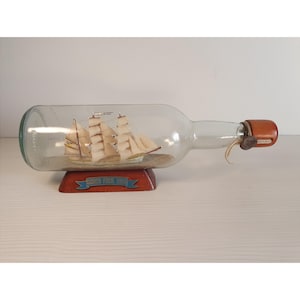 Puede incluir: Una botella de vidrio transparente que contiene un modelo de velero en miniatura. El modelo tiene velas beige y está montado sobre un soporte de madera. El soporte tiene una pancarta azul con el texto "GORCH FOCK 1958". La botella está sellada con un tapón marrón.