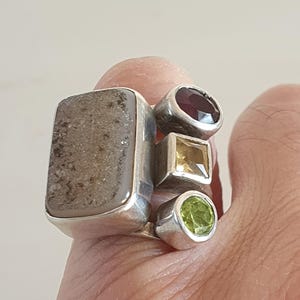 May include: A silver ring featuring multiple gemstones. The ring has a large, rectangular, gray stone, a round, deep red stone, a square, yellow stone, and a round, green stone. The gemstones are set in individual silver settings.