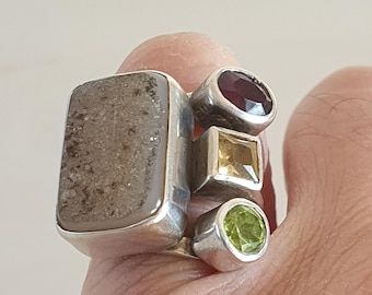 May include: A silver ring featuring multiple gemstones. The ring has a large, rectangular, gray stone, a round, deep red stone, a square, yellow stone, and a round, green stone. The gemstones are set in individual silver settings.