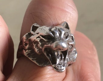 May include: A silver ring featuring a detailed wolf head design. The wolf's mouth is open, revealing its teeth. The ring is worn on a finger.