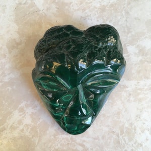 Könnte beinhalten: Ein grüner Malachit-Stein, der in die Form eines Gesichts geschnitzt ist. Das Gesicht hat ein detailliertes Design mit Augen, Nase und Mund.