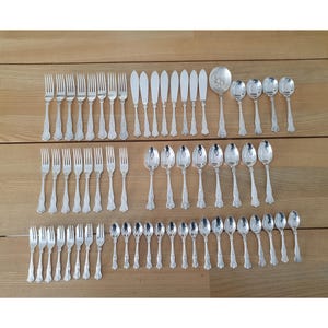 May include: A collection of vintage silverware, including forks, knives, and spoons, arranged on a wooden surface. The silverware has a shiny, silver finish and ornate handle detailing. The set includes various sizes of spoons and forks, perfect for a formal dining setting.