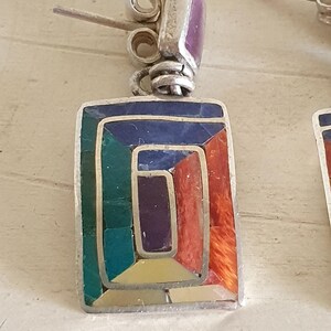 May include: A pair of silver earrings with a geometric design. The earrings are made of silver and feature a variety of colorful stones, including red, orange, yellow, green, blue, and purple.