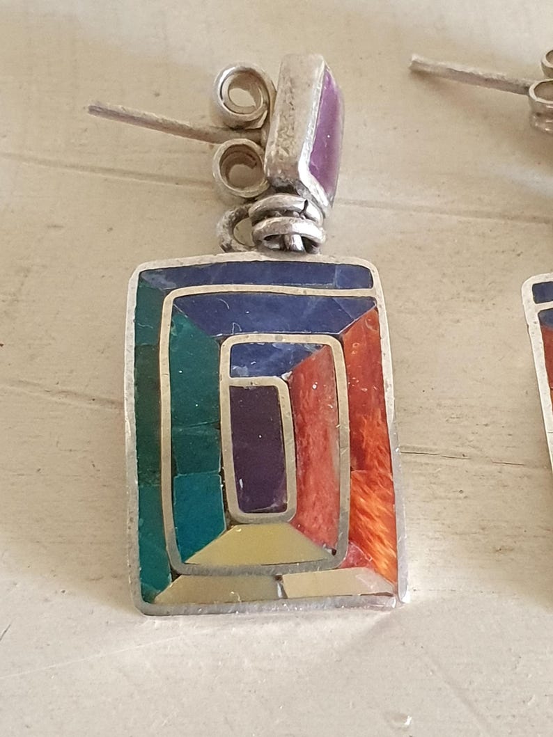 May include: A pair of silver earrings with a geometric design. The earrings are made of silver and feature a variety of colorful stones, including red, orange, yellow, green, blue, and purple.