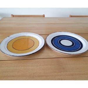 Könnte beinhalten: Zwei ovale Keramikplatten mit weißem Rand und strukturierter Kante. Eine Platte hat eine gelbe Mitte mit brauner Umrandung, die andere ein Design aus blauen und weißen konzentrischen Kreisen. Die Teller stehen auf einem Holztisch.