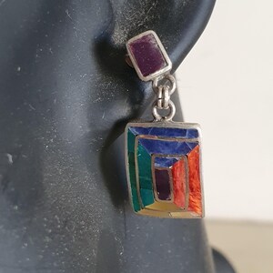 May include: A pair of silver dangle earrings with a rectangular shaped stone set in the center. The stone is made of multiple colors of inlaid stones, including green, blue, purple, orange, and yellow. The earrings have a small purple stone at the top.