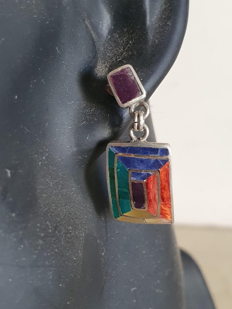 May include: A pair of silver dangle earrings with a rectangular shaped stone set in the center. The stone is made of multiple colors of inlaid stones, including green, blue, purple, orange, and yellow. The earrings have a small purple stone at the top.