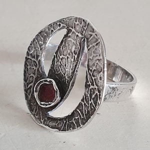 Op de afbeelding: Een zilveren ring met een getextureerd, ovaalvormig ontwerp en een kleine, ronde, rode steen. De ring heeft een unieke, artistieke uitstraling, met een ruwe, gehamerde metaaltextuur. De band is glad zilver.