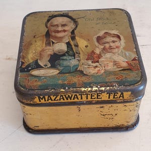 Puede incluir: Una caja de hojalata vintage con fondo dorado y un patrón floral. La tapa presenta una ilustración de dos personas, una mujer mayor y una niña más joven, ambas tomando té. El texto "Old folks at home" está impreso en la tapa. La parte inferior de la caja dice "Mazawatte Tea."