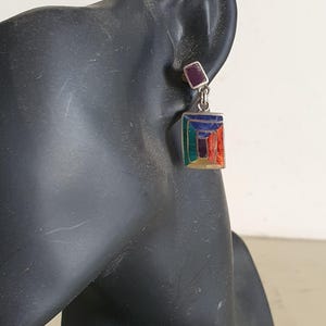 May include: Silver dangle earrings with a geometric design featuring multi-colored enamel. The top of the earring is a small square with a purple stone.