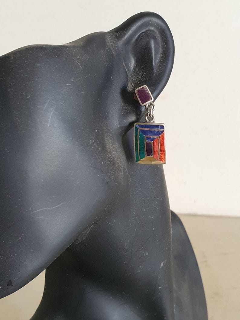 May include: Silver dangle earrings with a geometric design featuring multi-colored enamel. The top of the earring is a small square with a purple stone.